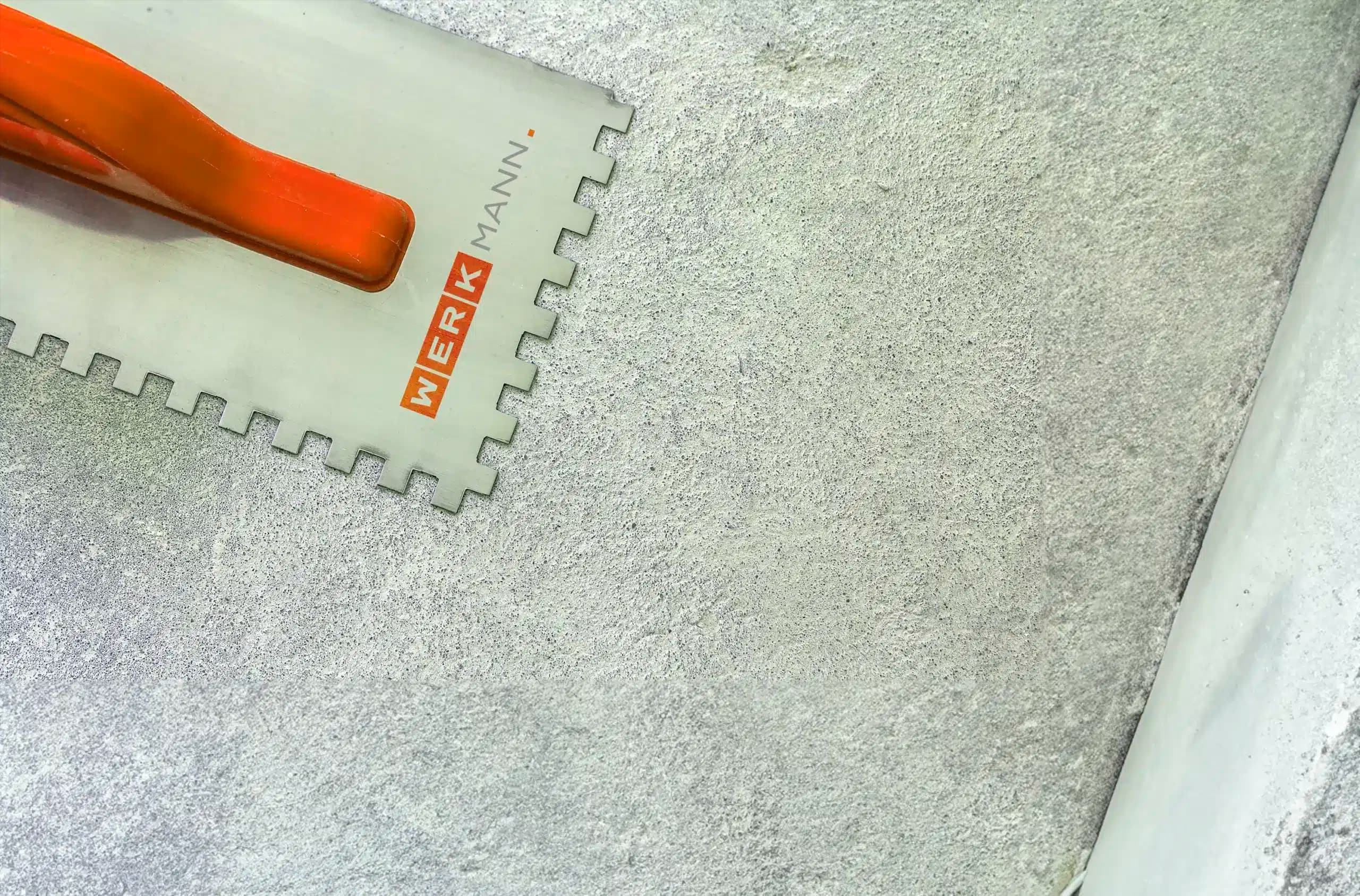 Eine Zahnkelle mit orangefarbenem Griff und der Aufschrift WERKMANN liegt auf einer rauen grauen Zementoberfläche, neben einer weißen Kante.