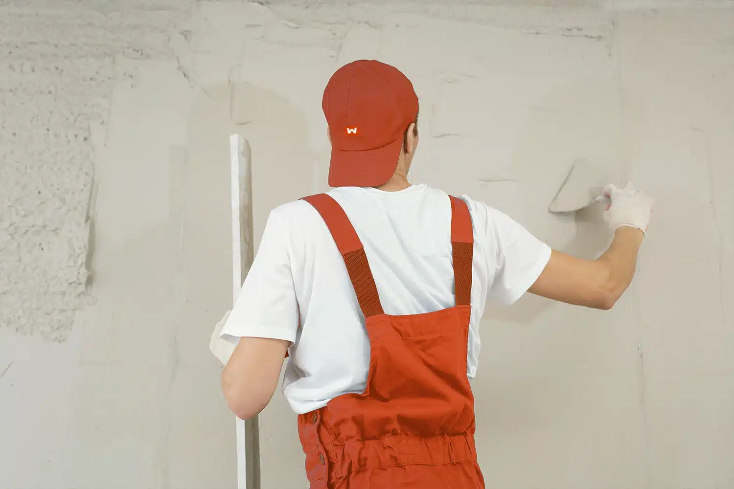Ein Handwerker in roter Latzhose und roter Werkmann-Kappe glättet mit der Kelle eine frisch verputzte Wand.