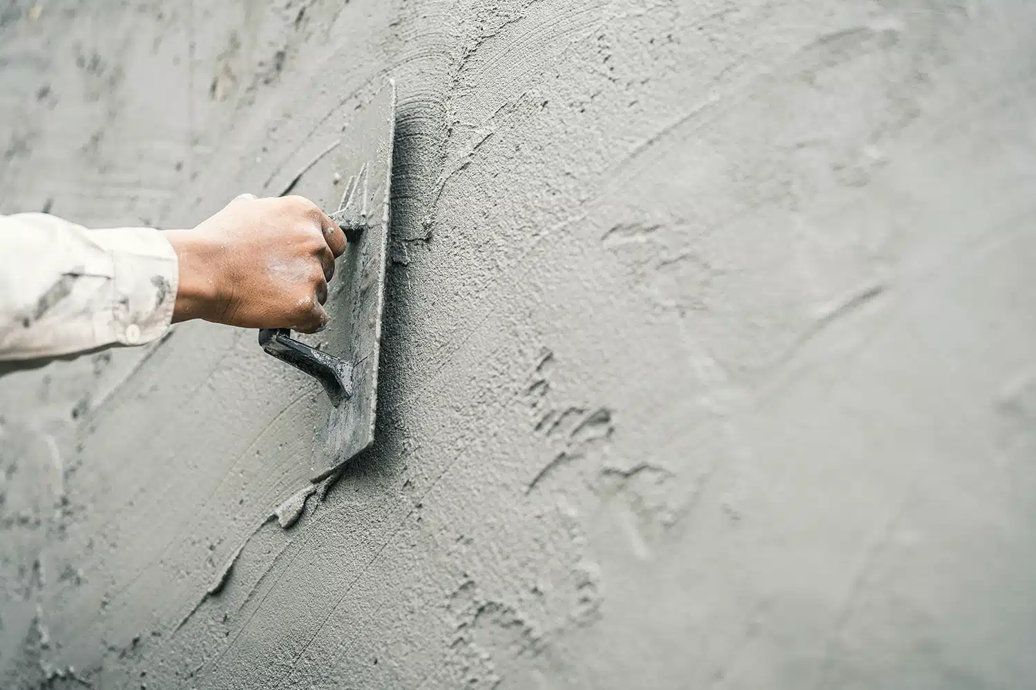 Eine Hand, die eine Kelle hält, glättet nassen Zement auf einer Wand und trägt eine gleichmäßige Putzschicht für Bau- oder Renovierungsarbeiten auf.