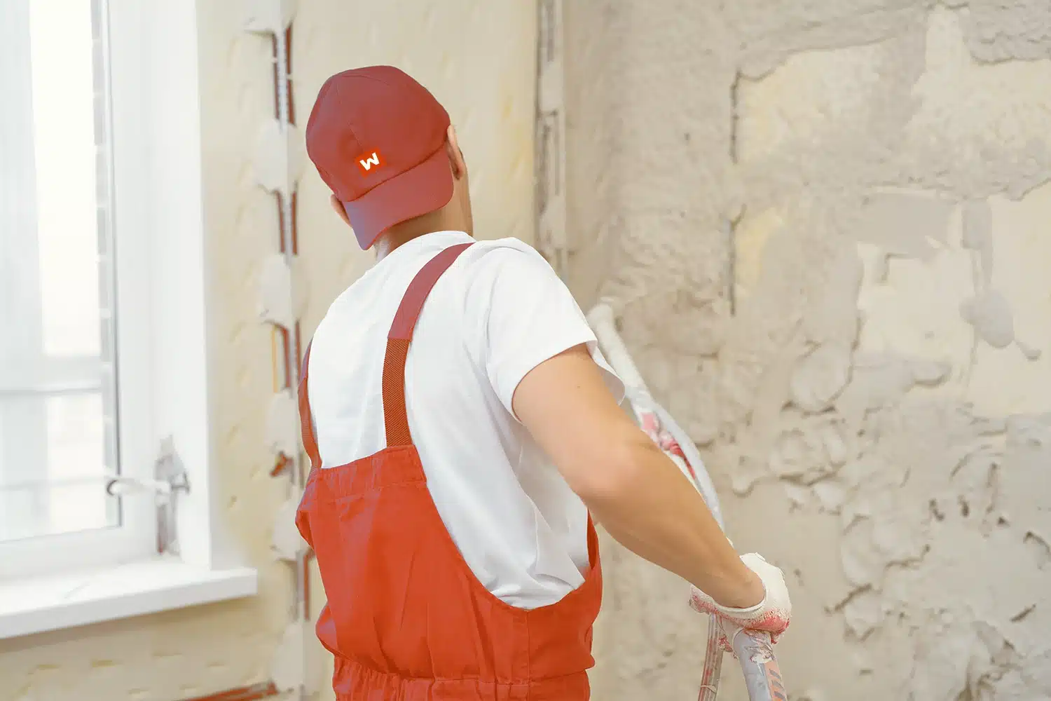 Eine Person mit roter Mütze, rotem Overall und weißen Handschuhen streicht oder verputzt eine Wand mit einer Rolle in einem Raum mit einem Fenster. Die Wand scheint teilweise entkleidet oder renoviert zu werden.