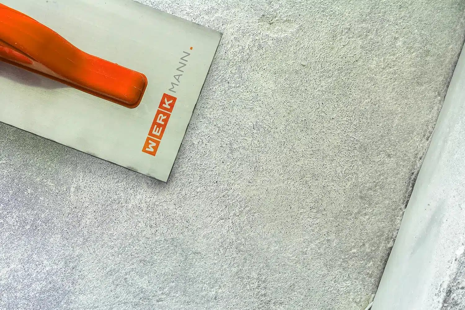 Nahaufnahme einer Metallkelle mit orangefarbenem Griff und dem Markennamen Werkmann, die auf einer glatten, grauen Betonfläche liegt.